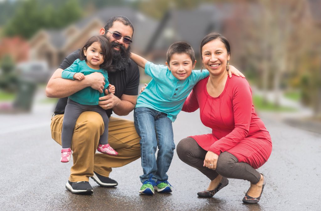 Smiling family with arms around each other: Mother, Father, pre-teen son and toddler daughter.